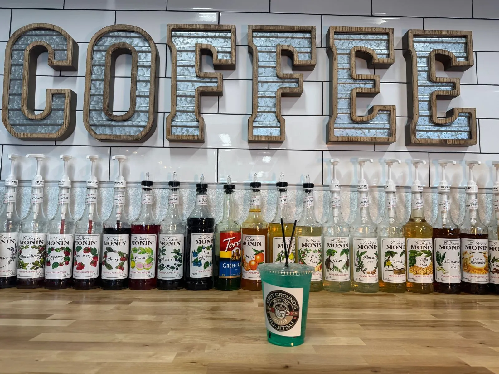 The coffee bar at Grit Grounds: a wall of Monin syrups under large COFFEE letters, with a green Lotus drink on the counter